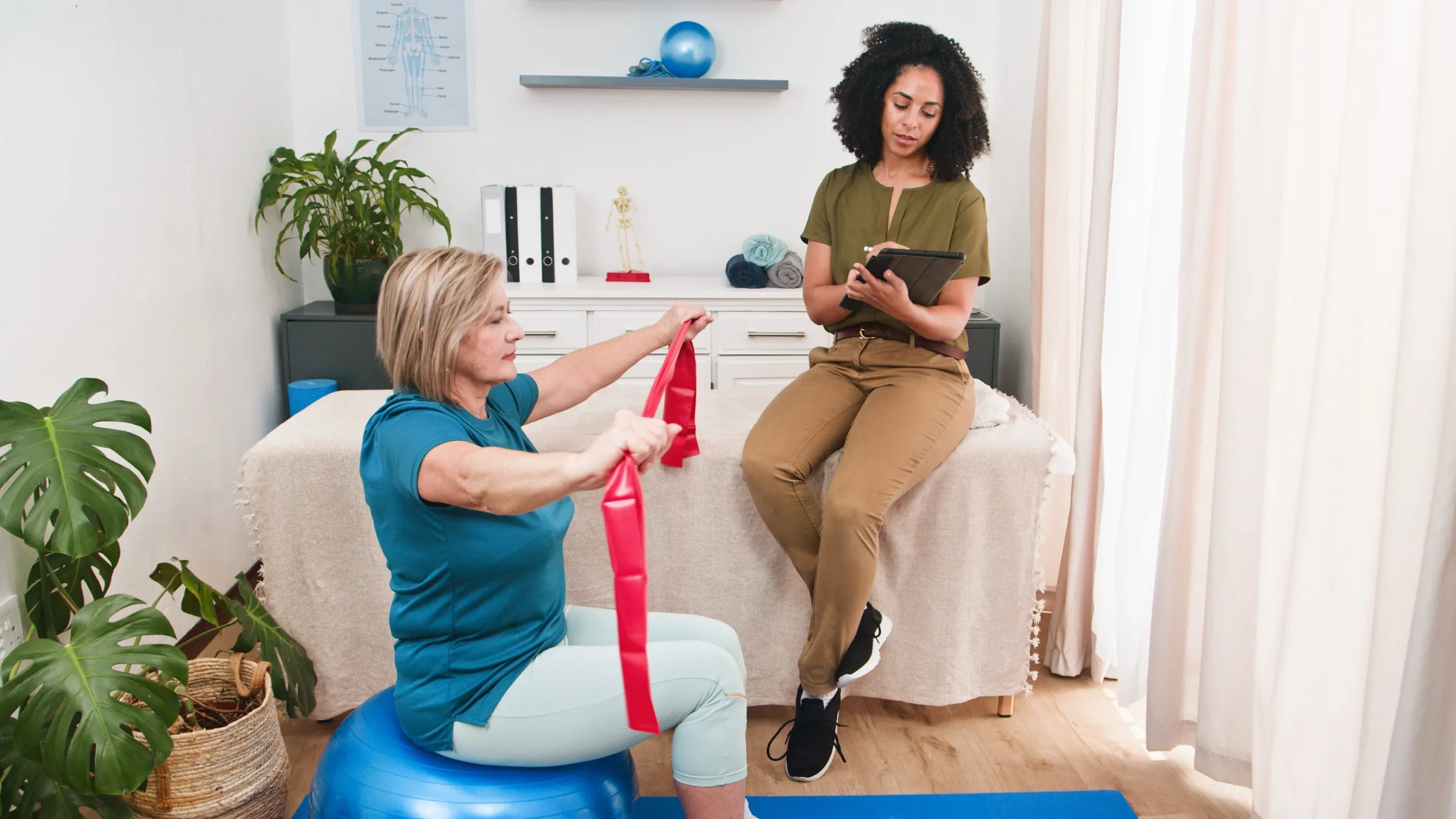A therapist taking notes on a tablet while an older adult exercises with a resistance band